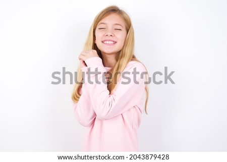 Dreamy beautiful caucasian little girl wearing pink sweater over white background with pleasant expression, closes eyes, keeps hands crossed near face, thinks about something pleasant