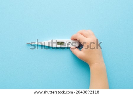 Baby hand holding white digital thermometer on light blue table background. Pastel color. Closeup. Temperature measuring of children. Healthcare concept. 