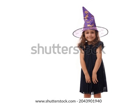 Portrait of pretty little girl wearing a wizard hat and dressed in stylish carnival dress, looking at camera posing with crossed arms against white background, copy space. Halloween concept