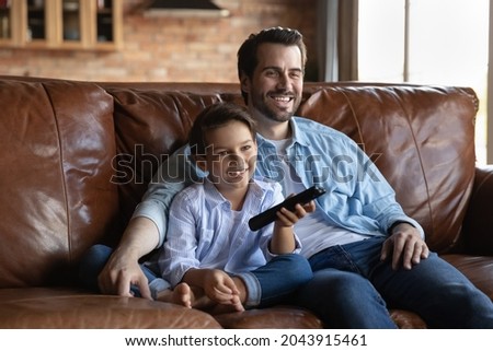Happy father with little son watching television show series or movie, relaxing sitting on cozy couch at home together, smiling 8s boy kid holding tv remote, switching channel, enjoying leisure time