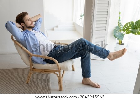 Side view untroubled millennial single man relaxing on chair in fashionable light apartment. Stress-free, daydreaming, comfort and modern house with climate control and air conditioner inside concept Royalty-Free Stock Photo #2043915563