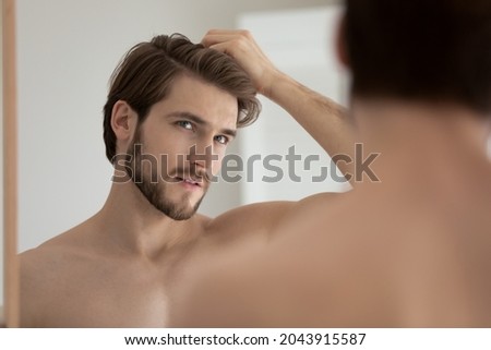 Young shirtless guy looks in mirror touch hair feels concerned due to receding, dry, dull hair. Hair loss common hair problem prevention, alopecia treatment ad, cosmetics products for male concept Royalty-Free Stock Photo #2043915587