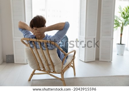 Rear view man relax on cozy chair with hands behind head daydreams looks into distance feels carefree enjoy weekend at modern home. No stress, day off leisure, comfort, air conditioner inside concept Royalty-Free Stock Photo #2043915596