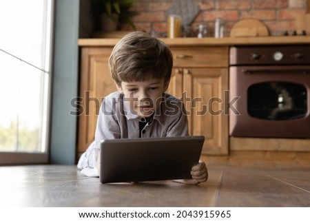 Adorable focused little preschool kid boy playing games on digital touchpad tablet, watching interesting photo video content online in social network, lying on warm floor in kitchen, tech addiction.