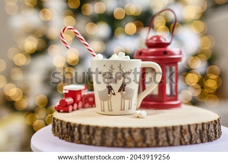 Fairy lights background, hot cocoa with marshmallows, white cup with deer on a wooden cut and red lantern and children's train at home during Christmas and New Year holidays. Soft focus bokeh.