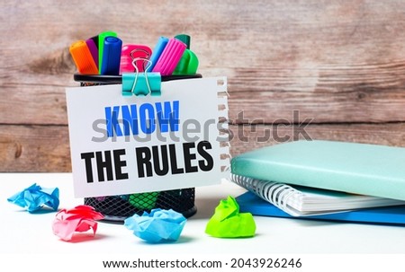 On the desktop and against the background of a wooden wall, there is a stand with multi-colored felt-tip pens, bright pieces of paper and a sheet of paper with the text KNOW THE RULES