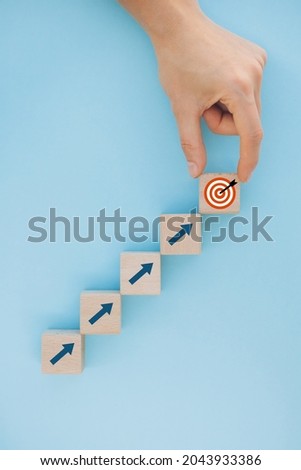 Close up of female hand arranging wood block stacking as step stair on blue background, putting down the cube with target icon on the top . Growth, success, strategy concept