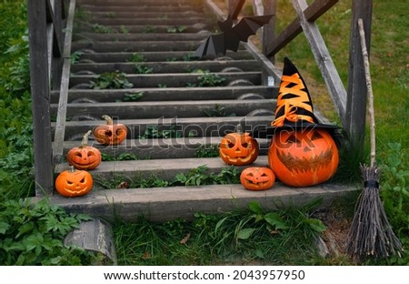 A large pumpkin with a witch's hat next to small pumpkins and a broom stand on the steps of the old staircase in front of the house. Jack o' lantern.  Halloween. trick or treating