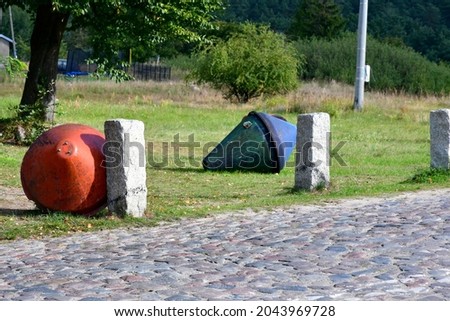 A close up on two safety buoys, one red and one green, laying next to some concrete posts next to a stone road with some shrubs and a dense forest seen behind them on a sunny summer day in Poland