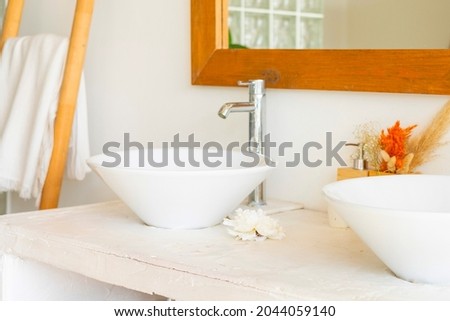 stylish and modern design bathroom interior with two white sinks, wicker baskets for cosmetics, mirror in wooden frame, dried flowers in vases, home plants in pot in Balinese style with white walls