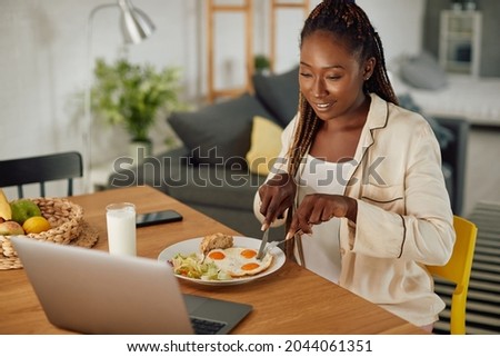 Young African American woman surfing the net on laptop while having breakfast at dining table.
