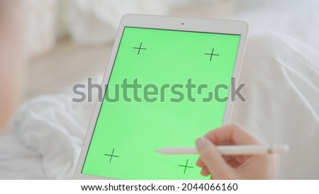 A tablet with a green screen for graphics. A woman with a beautiful pink manicure scrolls through the Internet pages on a tablet and puts likes. Uses a stylus, against the background of a white bed.