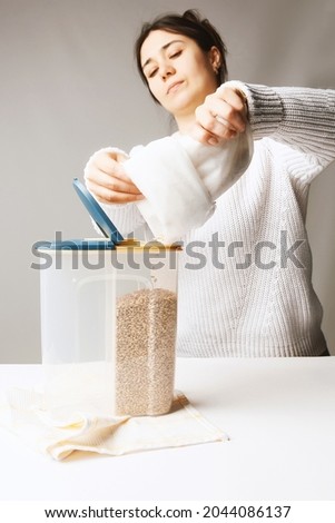 Shot of the girl pouring cereals into a two-compartment container. Plastic tableware is standing on the kitchen towel. Kitchen towel, white table, girl and container are on the gray background.
