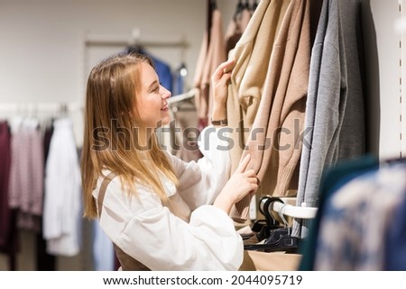 happy young woman girl chooses beige knitwear clothes for autumn or winter in clothing store. shopping in shop.