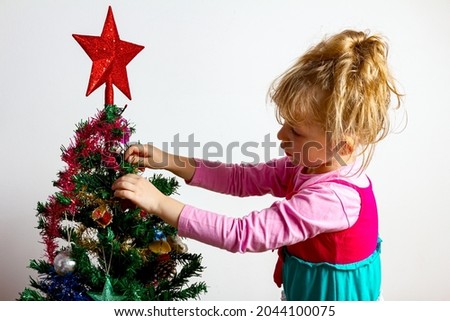 Preschooler female kid, child girl is placing shiny decorative strips with tassel on plastic evergreen, pine, tree for Christmas Eve.