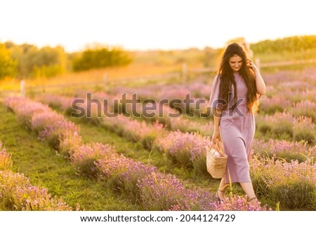 Young adult woman in stylish romantic dress walking at the lavender field with picnic basket. Beautiful brunette female spending weekends outdoors, enjoying fresh air, beautiful nature concept