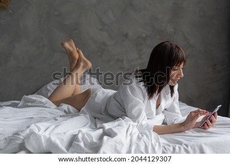 caucasian young woman in pajamas using phone lying in bed writing on social network in the morning, calm millennial woman relaxing on soft linen holding smartphone writing message. chatting at home