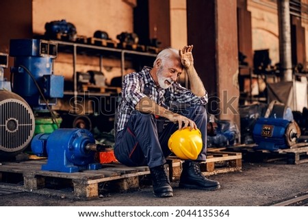 An old, tired bearded factory worker in overalls is sitting on the pallet and taking a break from hard work. He is wiping sweat from his forehead. Royalty-Free Stock Photo #2044135364