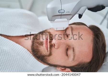 Bearded mustached dark-haired man dozing during a non-invasive face-lift procedure Royalty-Free Stock Photo #2044140047