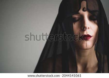 Redhead woman with a black veil on her head in a dark studio. A girl with an earring in her nose smiles slyly. Witch Makeup. Halloween costume.