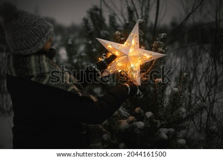 Merry Christmas! Woman putting big illuminated Christmas star on pine tree in snowy winter park at night. Magical Winter time. Stylish hipster female decorating christmas tree. Xmas miracle