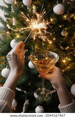 Happy New Year! Hands holding burning sparkler and champagne glass on background of christmas tree lights. Firework bengal light and drink in woman hands. Atmospheric moment. New Year eve party