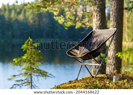 Camping folding chair with a book and a mug on the background of a forest lake. The chair is in focus, the background is blurred. Royalty-Free Stock Photo #2044168622