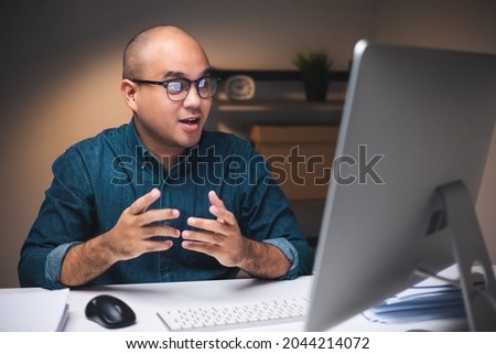 Young asian businessman working with computer video conference in the dark office at night. Attractive Indian man work hard overtime in home with floor lamp ambient warm light late at night