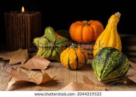 View of small pumpkins on green plaid blanket on wooden table with autumn leaves and burning candle, black background, horizontal