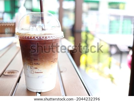 Ice coffee two layer of coffee and milk in closed plastic glass with straw on dark brown table and blur bright background in Thailand.
