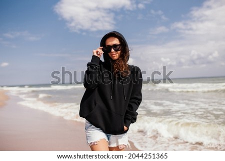Beautiful young adult woman in black hoodie or sweatshirt and sunglasses at the tropical beach. Mock-up for print. Space for your logo or design.