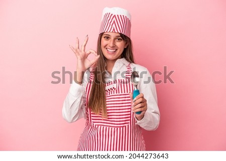 Young ice cream maker woman holding spoon isolated on pink background cheerful and confident showing ok gesture.