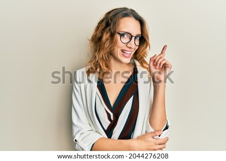 Young caucasian woman wearing business style and glasses with a big smile on face, pointing with hand and finger to the side looking at the camera. 