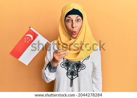 Middle age hispanic woman wearing hijab holding singapore flag scared and amazed with open mouth for surprise, disbelief face 