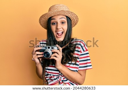 Young hispanic woman wearing summer hat holding vintage camera celebrating crazy and amazed for success with open eyes screaming excited. 
