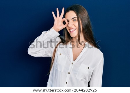 Young brunette teenager wearing casual white shirt smiling happy doing ok sign with hand on eye looking through fingers 
