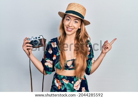 Young caucasian woman holding vintage camera smiling happy pointing with hand and finger to the side 