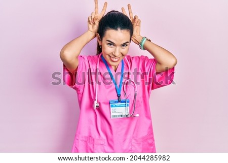 Young brunette woman wearing doctor uniform and stethoscope posing funny and crazy with fingers on head as bunny ears, smiling cheerful 
