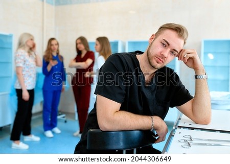 A photo of male medical professional sitting and smiling while his collegues are standing in the background.