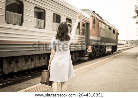 Young woman in with a suitcase waves to the departing train. Retro-style processing. The concept of being late for a train and seeing off or meeting a young person from a train on the platform. Royalty-Free Stock Photo #2044310351