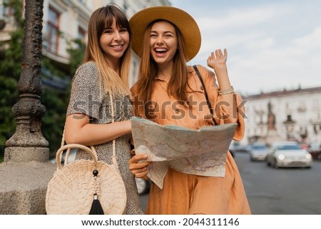 stylish young women traveling together in Europe dressed in spring trendy dresses and accessories smiling happy friends having fun taking photo on camera holding map