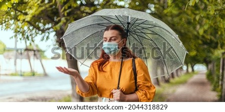 Unhappy woman in a protective medical mask standing under transparent umbrella and stretching hand catching water drops . Horizontal banner, copy space