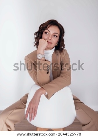 Beautiful pensive woman in stylish clothing sitting on white chair on white backdrop