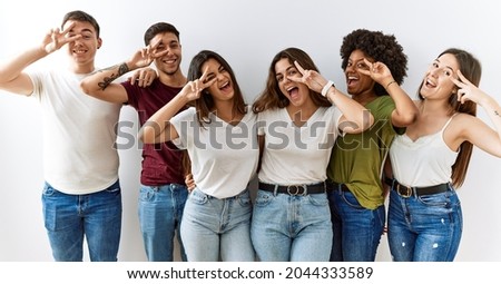 Group of young friends standing together over isolated background doing peace symbol with fingers over face, smiling cheerful showing victory 