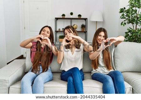 Group of three hispanic girls sitting on the sofa at home smiling in love doing heart symbol shape with hands. romantic concept. 