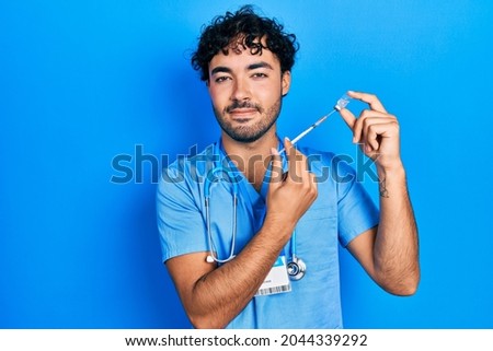 Young hispanic man wearing nurse uniform holding vaccine relaxed with serious expression on face. simple and natural looking at the camera. 