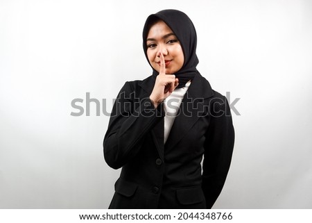 Beautiful asian young muslim business woman with finger on mouth, telling to be quiet, don't make noise, lower your voice, don't talk, isolated on white background