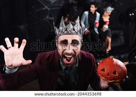 man in vampire king crown holding carved pumpkin and growling at camera during halloween party on black