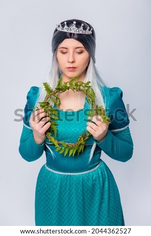 Portrait of a princess in a medieval, fantasy, turquoise dress with ash hair and a silver crown, posing with wreath of fern branches in her hands, isolated on a white background.