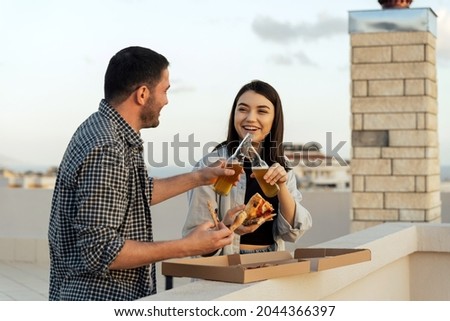 Couple enjoying cityscape and seaside view, drinking beer and eating pizza on the rooftop. Portrait of happy people spend quarantine time, picnic leisure on the roof on the sunset.